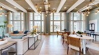 This single-floor open-concept gathering space embodies a refined coastal aesthetic, thoughtfully designed for both relaxation and socializing. Soaring ceilings finished with warm wood-plank paneling and crisp white structural beams define the architectural character of the room, while an eye-catching geometric chandelier with Edison-style bulbs serves as a dramatic centerpiece overhead.

The interior is bathed in natural light, courtesy of expansive floor-to-ceiling windows that span the full length of one wall, offering unobstructed views of an outdoor pool area and lush surroundings. Soft sage and seafoam-toned walls complement the light wood-look tile flooring throughout, reinforcing the coastal atmosphere.

The space is functionally divided into distinct zones. A comfortable lounge area anchors one end, featuring a linear tile-clad fireplace as its focal point. Adjacent to this is a dining area with a round table and woven rattan chairs. A long high-top bar counter with sleek black stools provides additional seating, connecting seamlessly to a built-in kitchenette. The kitchenette is finished with white subway tile, dark countertops, grey cabinetry, and stainless steel appliances, all illuminated by navy wall-mounted sconce lights. The overall ambiance is airy, welcoming, and elegantly curated.