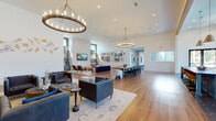 The Atwater Clubhouse in Oakwood, Georgia is a thoughtfully designed single-story community space that blends modern aesthetics with inviting, functional areas for residents to gather and relax. The open-concept floor plan flows seamlessly from one zone to the next, unified by warm hardwood flooring throughout and soaring ceilings accented by exposed wood beams in select areas.

At the heart of the space, a dramatic floor-to-ceiling stacked stone feature wall serves as a commanding architectural focal point, flanked by built-in shelving and accompanied by a curated lounge area with tufted leather seating arranged on a patterned area rug. Oversized circular chandelier fixtures with exposed Edison bulbs hang overhead, providing warm, ambient illumination alongside recessed lighting.

A generously sized communal dining area features a live-edge wood table surrounded by modern black chairs, ideal for community events or casual gatherings. Adjacent, a stylishly appointed kitchen counter with a bold dark surface, teal cabinetry, white subway tile backsplash, and leather-upholstered bar stools opens toward the exterior, bathed in natural light from large windows with views of the surrounding property.

Additional seating nooks with built-in benches, decorative wallcoverings, and curated wall art create intimate pockets within the larger space. The clubhouse's design strikes a balance between refined style and comfortable community living.