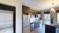This contemporary single-floor residence showcases a thoughtfully designed interior that blends style and functionality. The kitchen stands as a highlight of the space, featuring a streamlined galley-style layout with rich, dark espresso cabinetry extending to the ceiling for ample storage. Stainless steel appliances — including a full-size refrigerator, dishwasher, electric range, and built-in microwave — lend a polished, modern aesthetic. The countertops are complemented by a classic tile backsplash, while an undermount sink and modern faucet add refined detail. Track lighting runs along the ceiling, providing focused illumination across the workspace. Warm hardwood floors flow throughout, anchoring the space with a sense of warmth and continuity. A window near the kitchen allows natural light to filter in, offering a glimpse of greenery outside and enhancing the overall brightness of the interior. An open shelving unit provides additional display and storage options. The overall ambiance is clean, modern, and inviting, with a cohesive design palette of dark cabinetry against neutral walls creating a striking yet comfortable environment.