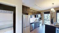 This contemporary single-floor residence showcases a thoughtfully designed interior that blends style and functionality. The kitchen stands as a highlight of the space, featuring a streamlined galley-style layout with rich, dark espresso cabinetry extending to the ceiling for ample storage. Stainless steel appliances — including a full-size refrigerator, dishwasher, electric range, and built-in microwave — lend a polished, modern aesthetic. The countertops are complemented by a classic tile backsplash, while an undermount sink and modern faucet add refined detail. Track lighting runs along the ceiling, providing focused illumination across the workspace. Warm hardwood floors flow throughout, anchoring the space with a sense of warmth and continuity. A window near the kitchen allows natural light to filter in, offering a glimpse of greenery outside and enhancing the overall brightness of the interior. An open shelving unit provides additional display and storage options. The overall ambiance is clean, modern, and inviting, with a cohesive design palette of dark cabinetry against neutral walls creating a striking yet comfortable environment.