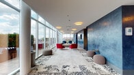 Located at 1000 S Clark St in Chicago's South Loop neighborhood, this rooftop amenity floor offers a sophisticated blend of social and functional spaces designed for community living. The centerpiece is a generously proportioned lounge lined with floor-to-ceiling glass walls that flood the interior with natural light and frame sweeping panoramic views of the Chicago skyline and Lake Michigan. Vibrant red and navy accent chairs, a neutral sofa, and patterned area rugs create a stylish, inviting atmosphere throughout the lounge. Direct access to a furnished outdoor terrace provides an extension of the living space, complete with striped lounge seating and large planters against a stunning urban backdrop. Adjacent to the lounge, a fully equipped demonstration kitchen stands out with its navy-tiled island, quartz countertops, subway tile backsplash, stainless steel appliances, a commercial-grade range and hood, and chalkboard accent walls. A dedicated dining area with a long communal table, pendant lighting, and a wall-mounted TV offers an ideal setting for group gatherings. Rich blue textured walls and patterned tile flooring tie the design together cohesively, making this a compelling and well-appointed shared amenity space.
