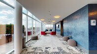 Located at 1000 S Clark St in Chicago's South Loop neighborhood, this rooftop amenity floor offers a sophisticated blend of social and functional spaces designed for community living. The centerpiece is a generously proportioned lounge lined with floor-to-ceiling glass walls that flood the interior with natural light and frame sweeping panoramic views of the Chicago skyline and Lake Michigan. Vibrant red and navy accent chairs, a neutral sofa, and patterned area rugs create a stylish, inviting atmosphere throughout the lounge. Direct access to a furnished outdoor terrace provides an extension of the living space, complete with striped lounge seating and large planters against a stunning urban backdrop. Adjacent to the lounge, a fully equipped demonstration kitchen stands out with its navy-tiled island, quartz countertops, subway tile backsplash, stainless steel appliances, a commercial-grade range and hood, and chalkboard accent walls. A dedicated dining area with a long communal table, pendant lighting, and a wall-mounted TV offers an ideal setting for group gatherings. Rich blue textured walls and patterned tile flooring tie the design together cohesively, making this a compelling and well-appointed shared amenity space.