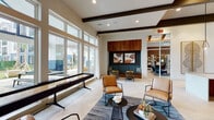 This single-floor amenity space is designed with a contemporary aesthetic, blending warm wood accents, light tile flooring in a herringbone pattern, and bold dark exposed ceiling beams throughout. The open layout is organized into several distinct yet connected zones, creating a versatile environment for socializing, recreation, and relaxation.

The main lounge area features an inviting arrangement of tan leather chairs, low-profile coffee tables, and layered area rugs, complemented by curated artwork. A striking wood-paneled fireplace wall with integrated lighting serves as a focal point, while a separate seating nook with a tufted white sofa and gallery wall arrangement offers a more intimate setting.

The games area is well-equipped with a foosball table and shuffleboard, bathed in natural light from floor-to-ceiling windows that frame views of an outdoor courtyard with additional seating. A spacious bar and lounge counter features a dramatic marble waterfall countertop, warm wood cabinetry, pendant lighting, and a built-in refreshment station with a mounted television. An adjacent co-working or meeting room is visible through glass partitions, completing this multifunctional amenity space.