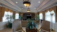 This refined single-floor space exudes classic elegance through its thoughtfully designed interior. The living area immediately draws attention with its distinctive tray ceiling, adorned with crisp crown molding that frames the room with architectural sophistication. Two ornate wrought-iron pendant light fixtures with amber glass shades serve as striking focal points, casting a warm glow throughout the space.

The room centers around a built-in entertainment unit crafted in rich dark wood, flanked by decorative shelving and cabinetry that add both function and visual depth. Textured, faux-finished walls in warm earth tones provide a backdrop that enhances the traditional aesthetic. Generous windows dressed with layered valances and draped curtains allow ample natural light to fill the space while maintaining an air of refinement.

The seating arrangement features plush, neutral-toned upholstered furniture, including classic armchairs with gilded wooden frames, inviting relaxed comfort. A decorative side table hosts a lush tropical plant arrangement, reinforcing the space's connection to natural elements. Light-toned tile flooring ties the room together, complementing the overall warm and inviting palette. The result is a space that balances grandeur with comfort.
