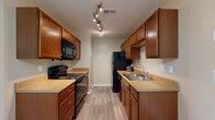 This single-floor residence offers a thoughtfully designed layout that balances functionality with comfort throughout. The galley-style kitchen is a standout feature, lined on both sides with rich, warm-toned wood cabinetry and complemented by light stone-finish countertops. Black appliances — including a range, over-the-range microwave, refrigerator, and dishwasher — provide a cohesive look, while wood-look plank flooring adds durability and visual appeal. Distinctive track lighting overhead ensures excellent task illumination.

The living area is generously proportioned, featuring vaulted ceilings with a ceiling fan and a sliding glass door that invites natural light and opens to an outdoor space. Plush carpet extends throughout the living, dining, and bedroom areas, creating a warm and comfortable feel. The dining area is defined by an elegant chandelier, seamlessly connecting to the main living space.

The bedroom is well-sized, brightened by a bay-style window arrangement that frames views of greenery outside. Neutral beige walls throughout the residence create a calm, cohesive atmosphere. A utility area with washer/dryer hookups and a vanity with matching cabinetry and countertops rounds out this practical and inviting space.
