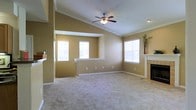 This single-story space showcases an open-concept living area characterized by vaulted ceilings that lend a sense of spaciousness and airiness to the interior. The living room is anchored by a classic white-mantled fireplace on one wall, providing a natural focal point and a cozy atmosphere. A ceiling fan with an integrated light fixture is centrally mounted, adding both functionality and a finishing architectural detail to the soaring ceiling.

The flooring throughout the main living area consists of neutral-toned carpet, contributing to a warm and comfortable feel underfoot. Walls are painted in a warm tan hue, complementing the light-colored crown molding and white baseboards that frame the space with a clean, traditional aesthetic.

Multiple windows are strategically positioned to maximize natural light, brightening the interior and creating an inviting ambiance. A partial wall with a pass-through opening connects the living area to an adjacent space, likely a kitchen or dining area, enhancing the open flow of the floor plan. The overall design reflects a practical and approachable layout well-suited for everyday living.