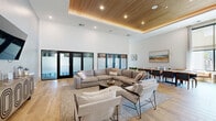 This thoughtfully designed single-floor space offers a generous, open-concept layout that blends sophisticated style with functional versatility. The interior immediately impresses with its soaring ceilings, which amplify the sense of volume throughout. A standout architectural feature is the coffered tray ceiling finished in warm slatted wood planks, fitted with recessed lighting that casts an inviting, even glow across the space.

Light hardwood flooring extends seamlessly throughout, complementing the bright, white walls and contributing to an airy, contemporary aesthetic. The main area is organized into distinct zones: a comfortable lounge area furnished with a large sectional sofa, accent chairs, and a round coffee table, ideal for casual gatherings; and a formal dining zone anchored by an elongated table with teal upholstered chairs, suited for group meals or meetings.

A wall-mounted television and additional seating area on the far end offer a media-focused retreat. Tall frosted glass panels with sleek black frames separate an adjoining room, allowing privacy while maintaining visual openness. Large windows invite natural light and outdoor views, enhancing the welcoming atmosphere. A hallway with curated wall art and direct exterior door access provides a clean, functional entry and exit point.