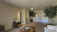 This single-floor residence offers a comfortable and well-organized layout designed for easy everyday living. The main living area is spacious and welcoming, featuring neutral beige walls and plush wall-to-wall carpeting that lend a warm, understated tone throughout. Flush ceiling lighting keeps the space bright and well-illuminated, while decorative touches such as a large potted indoor tree add a natural, refreshing element to the interior.

The open layout allows for a smooth transition from the living room toward the kitchen area, which is visible in the background and benefits from natural light streaming through what appears to be a sliding glass door or large window. The kitchen area features cabinetry and bright, airy finishes that complement the rest of the home.

Multiple doorways branch off from the main living space, leading to additional rooms including what appears to be a bathroom and other private areas, suggesting a practical and efficient floor plan. A dedicated entry door provides clear separation between the interior and exterior of the home.

Overall, the space presents a clean, move-in-ready aesthetic with a calm, neutral palette, functional room connectivity, and a layout that balances open communal areas with more private spaces — making it a versatile and livable environment.