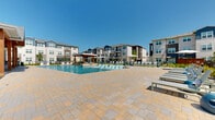 This contemporary residential community is centered around a striking outdoor amenity area designed for both relaxation and recreation. The focal point is a generously sized resort-style swimming pool with crystal-clear blue water, featuring a wading ledge with in-pool lounge chairs, and a separate splash zone. The pool is surrounded by an expansive stone paver deck that provides ample space for sunbathing and socializing.

A covered outdoor pavilion anchors one end of the pool area, framed by warm wood timber beams and a pitched roof, offering a shaded gathering space adjacent to the main clubhouse building. The clubhouse itself features large floor-to-ceiling windows and a covered porch with outdoor seating. On the opposite side of the pool, a pergola-covered grill and lounge station provides an additional outdoor entertaining hub.

Rows of modern sling-style chaise lounges are arranged throughout the deck, accompanied by umbrella-shaded seating areas. The pool zone is enclosed by sleek black metal fencing, beyond which a well-maintained grassy lawn provides open green space ideal for outdoor games, including cornhole. The surrounding multi-story residential buildings feature contemporary architecture with a mix of board-and-batten siding, balconies, and metal roof accents, creating a cohesive and vibrant community atmosphere.