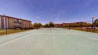Located at 201 Idlewild Rd in Bel Air, Maryland, this outdoor recreational facility features a set of hard-surface tennis courts finished in a classic green playing surface with crisp white and yellow boundary line markings. The courts are fully enclosed by sturdy chain-link fencing, providing both safety and clear boundary definition for players. A regulation net divides the primary court, making the space ready for competitive or recreational play.

The courts are situated within a larger residential community, surrounded by multi-story brick residential buildings that feature balconies and are set against lush, well-maintained green lawns. Mature trees line the perimeter, offering shade and a pleasant natural backdrop. Views from the courts extend to a nearby community pool area and clubhouse, highlighting the broader array of amenities available on the grounds.

The open-air setting benefits from abundant natural light and clear sightlines across the complex. The courts show visible signs of weathering and surface wear, indicating established use over time. Lamp posts positioned near the fencing suggest the possibility of extended play into the evening hours. Overall, the space serves as a central outdoor amenity within the Idlewild Road community, offering residents a dedicated venue for tennis and active recreation in a green, open-air environment.