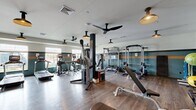 Located at Kingston Collection in Kingston, Massachusetts, this fitness center offers a comprehensively equipped workout facility on a single floor. The space is thoughtfully designed with a distinctive aesthetic, featuring teal and white walls accented by a bold orange horizontal stripe, dark hardwood-style flooring, and a mix of industrial-style pendant lighting and ceiling fans that contribute to both comfort and ambiance.

Natural light flows in through a row of large windows along one wall, overlooking the surrounding exterior and lending the space an open, airy feel. The cardio zone is well-appointed with treadmills, elliptical machines, and a rowing machine positioned near the windows for an energizing workout experience. The strength training area is equally robust, featuring a power rack, a dual cable crossover machine, a RaX functional training system with integrated display, FreeMotion cable machines, adjustable weight benches, and a full dumbbell rack with free weights ranging across various sizes. Medicine balls and plyo boxes round out the functional fitness offerings. Mirrored wall sections enhance the sense of space and support proper form during training. The layout is open and well-organized, allowing for comfortable movement throughout the facility.