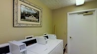 Located at 5680 Hibernia Dr in Columbus, Ohio, this community laundry facility offers residents a practical and well-maintained space for their everyday laundry needs. The single-floor layout is straightforward and functional, designed with convenience in mind. The room features multiple commercial-grade Speed Queen washing machines lined up along the wall, providing reliable, high-capacity cleaning performance suited for regular residential use. The walls are finished in a warm, neutral green tone, complemented by a textured ceiling that adds a subtle visual detail to the space. A decorative framed print of the Hibernia community signage adorns the wall, adding a welcoming, community-oriented touch to an otherwise utilitarian room. Natural light and clean white trim around the doorway contribute to the bright, tidy atmosphere of the facility. The space is neatly organized, ensuring ease of access and movement throughout. This laundry room serves as a shared community amenity, reflecting the Hibernia community's commitment to providing practical, well-kept facilities for its residents in the Columbus area.