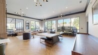 Located at 50 Estuary Trail in Alpharetta, GA 30005, this 770-square-foot single-floor community amenity space is thoughtfully designed to blend sophisticated style with functional recreational use. The open-concept layout is anchored by a striking herringbone-patterned hardwood floor that flows seamlessly throughout both zones of the space.

The lounge area is defined by floor-to-ceiling black-framed windows that flood the interior with abundant natural light and offer views of the surrounding greenery and outdoor amenities. A large, channeled sectional sofa in a warm neutral tone anchors the seating arrangement, complemented by a textured blue accent chair and a live-edge wood coffee table. A built-in wood shelving unit with a sliding barn door showcases a dramatic abstract artwork, adding a bold artistic focal point. Contemporary sputnik-style chandeliers provide stylish overhead lighting throughout.

Adjacent to the lounge, the game area features a prominent ping-pong table with sleek upholstered seating, alongside a solid wood shuffleboard table. Large windows and direct outdoor access enhance the airy atmosphere of this zone. Dark crown molding frames the crisp white walls, lending a polished, upscale character to the entire space.