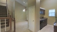This two-story residence showcases a thoughtfully designed interior that emphasizes vertical space and an open layout. The main living area is a standout feature, boasting dramatically vaulted ceilings that create an expansive, airy atmosphere. A pendant light hangs centrally, drawing the eye upward and adding a decorative focal point to the room. Neutral-toned carpet and warm beige walls contribute to a cohesive, understated aesthetic throughout the space.

Adjacent to the living area, the kitchen is outfitted with dark espresso-toned cabinetry that provides a rich contrast against the lighter surroundings. A dark countertop with an integrated sink and black appliances completes the functional workspace, offering ample storage and prep space.

A paneled interior door provides access to an additional room or closet, maintaining a clean and classic architectural style consistent throughout the home. A staircase with a dark wood newel post connects the main level to the upper floor, adding a traditional design element. The overall ambiance is calm and neutral, offering a versatile backdrop suitable for a variety of interior styles.