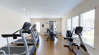 Located at 100 Durham Park Way in Pooler, Georgia, this fitness center offers a well-appointed, single-floor exercise facility designed to support a comprehensive workout routine. The space features warm hardwood flooring throughout, which adds a clean and polished aesthetic to the room while providing a durable surface suited for active use. Natural light flows in through multiple windows fitted with blinds, creating a bright and energizing atmosphere that enhances the workout environment. The layout is thoughtfully organized to accommodate a variety of exercise equipment without feeling cramped. A row of treadmills lines one side of the room, providing ample cardio options for residents. A recumbent exercise bike is positioned along the opposite side, offering a low-impact cardiovascular alternative. At the center of the space stands a multi-station cable and weight machine, enabling a wide range of strength training exercises. Recessed lighting in the smooth white ceiling supplements the natural light, ensuring the room remains well-lit at any time of day. The neutral, light-toned walls contribute to an open and clean ambiance, making the space feel welcoming and functional. Overall, this fitness center provides a practical and comfortable environment for residents looking to maintain an active lifestyle.