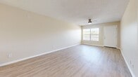 Located at 5290 70th Ave N in Pinellas Park, Florida, this 2-bedroom, 1-bathroom unit offers a thoughtfully laid-out single-floor plan with a clean, updated aesthetic throughout. Light wood-style laminate flooring runs continuously through the living areas, bedrooms, and hallway, creating a cohesive and easy-to-maintain finish. The open living room is bright and airy, featuring a ceiling fan with light fixture, a textured ceiling, and a large window that invites natural light and outdoor views of the surrounding landscape. The kitchen is well-appointed with white shaker-style cabinetry, granite-look countertops, black appliances including a refrigerator and electric range, and a window above the sink. Both bedrooms are comfortably proportioned, each with ceiling fans and windows that bring in natural light. The primary bedroom includes a reach-in closet with shelving. The shared bathroom features a tub-shower combination with white tile surround, a white vanity with granite-look countertop, and a curtain rod. A connecting hallway links the bedrooms, bathroom, and living areas efficiently. The building exterior features balconies, mature trees, and a pool area visible from the grounds.