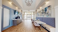 Located at 4910 Samaritan Way in Jacksonville, FL, this single-floor community clubhouse is thoughtfully designed to offer a range of social and recreational amenities. The welcoming lobby greets visitors with soaring tray ceilings, an elegant multi-tiered chandelier, warm hardwood-style flooring, and French doors that flood the space with natural light and provide views to an outdoor terrace. A service credenza finished in a calming blue palette, paired with wall-mounted displays and contemporary artwork, gives the entry an organized, polished feel. Built-in booth seating with marble-topped tables and upholstered banquettes offers comfortable casual dining, accented by stylish pendant lighting and a textured striped feature wall. The lounge area features coffered ceilings with exposed dark wood beams, a dramatic sputnik chandelier, and expansive windows that connect the interior to the outdoor patio. Plush sectional seating and layered rugs contribute to a relaxed yet refined atmosphere. Adjacent to the lounge, a game room houses a full-size pool table, bar-height seating, and direct patio access through French doors. The kitchen area is a standout, showcasing shaker-style cabinetry, a shimmering blue glass tile backsplash, quartz countertops, open wood shelving, stainless steel appliances, and a striking live-edge communal dining table — ideal for gatherings and events.