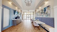 Located at 4910 Samaritan Way in Jacksonville, FL, this single-floor community clubhouse is thoughtfully designed to offer a range of social and recreational amenities. The welcoming lobby greets visitors with soaring tray ceilings, an elegant multi-tiered chandelier, warm hardwood-style flooring, and French doors that flood the space with natural light and provide views to an outdoor terrace. A service credenza finished in a calming blue palette, paired with wall-mounted displays and contemporary artwork, gives the entry an organized, polished feel. Built-in booth seating with marble-topped tables and upholstered banquettes offers comfortable casual dining, accented by stylish pendant lighting and a textured striped feature wall. The lounge area features coffered ceilings with exposed dark wood beams, a dramatic sputnik chandelier, and expansive windows that connect the interior to the outdoor patio. Plush sectional seating and layered rugs contribute to a relaxed yet refined atmosphere. Adjacent to the lounge, a game room houses a full-size pool table, bar-height seating, and direct patio access through French doors. The kitchen area is a standout, showcasing shaker-style cabinetry, a shimmering blue glass tile backsplash, quartz countertops, open wood shelving, stainless steel appliances, and a striking live-edge communal dining table — ideal for gatherings and events.