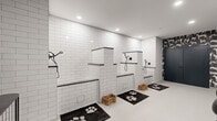 Located at 3111 W Allegheny Ave in Philadelphia, PA, this purpose-built pet grooming space combines striking aesthetics with practical functionality. The single-floor layout is thoughtfully organized to accommodate multiple grooming stations along one wall, each featuring raised tiled platforms with dark-trimmed edges, built-in matte black handheld shower fixtures, and woven storage baskets beneath. The walls of the grooming area are clad floor-to-ceiling in classic white subway tile, lending a clean, hygienic, and polished look throughout. Recessed ceiling lighting illuminates the space evenly, enhancing the bright, open feel. A dramatic design transition occurs toward the entrance area, where bold black wallpaper featuring an illustrated pattern of various dog breeds creates a visually striking focal point. This section also includes a set of teal double doors, a sculptural black balloon-dog accent piece, and a sleek console display with canine-themed artwork. Black paw print rugs are positioned at each grooming station, tying the playful theme together cohesively. The smooth white flooring flows seamlessly throughout, unifying both zones of the space. The overall ambiance is equal parts functional grooming facility and thoughtfully curated boutique environment, purpose-designed for pet care professionals and their clients.
