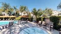 This outdoor amenity area is a thoughtfully designed recreational space that offers residents a resort-style experience. At its center sits a generously sized swimming pool alongside a bubbling spa, both maintained to a high standard and surrounded by a clean, spacious concrete deck. Multiple sling-style lounge chairs are arranged throughout the deck, providing ample seating for sunbathing and relaxation, while large cantilever umbrellas offer shaded retreats from the sun.

The space is framed by mature, towering palm trees and well-manicured hedgerows that create a sense of privacy and tropical ambiance. Additional seating areas with chairs are nestled among the greenery near the spa, offering quieter spots to unwind. Clearly posted spa rules and safety signage reflect attentive property management.

A tiled outdoor shower station with striped grey-and-white wall tiles and a fold-down bench is conveniently located adjacent to the pool area, allowing for a quick rinse. A key-access restroom facility is also available on-site for pool area guests. Wide, accessible concrete pathways connect all areas of the space, bordered by neatly landscaped shrubs and trees. The surrounding multi-story residential buildings with tile rooflines and balconies are visible, enhancing the cohesive community feel of this outdoor retreat.