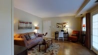 This single-floor space offers a thoughtfully designed open layout that seamlessly connects the living, dining, and workspace areas. The living room serves as the central hub, featuring warm neutral-toned walls that create a welcoming ambiance. A plush sofa anchors the seating area, complemented by a floral-patterned area rug, a wooden coffee table, and decorative artwork that adds personality to the space. A flat-screen television is mounted on a dark media console, providing a natural focal point for the room. Adjacent to the living area, a cozy dining nook accommodates a bistro-style table and chairs beneath a ceiling fan, with a window dressed in warm curtains that allows natural light to filter in. A compact yet functional workspace is tucked neatly into the living area, featuring a sleek desk, chair, and accent lamp — ideal for work or study. The flooring transitions between tile and hardwood-style surfaces, adding visual interest and texture throughout. Soft, layered lighting from floor lamps and table lamps enhances the warm, residential atmosphere. The space is efficiently organized, making excellent use of its single-level footprint while maintaining a comfortable and livable feel.