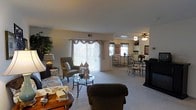This single-story residence features an open-concept layout that seamlessly connects the living and dining areas, creating a spacious and functional flow. The living area is carpeted and comfortably accommodates a variety of seating arrangements, including a sofa, accent chair, and a glass-topped coffee table. A large sliding glass door with vertical blinds and decorative valance allows natural light to fill the space while providing access to the outdoors. Adjacent to the living area, the dining space features an elegant wrought-iron table and chair set, positioned beneath a ceiling fan with a light fixture that provides ambient overhead lighting. A mirror and framed artwork on the walls add decorative character to the dining area. The kitchen is partially visible beyond the dining space, offering a glimpse of additional cabinetry and countertop space. Throughout the residence, the neutral color palette of beige walls and light carpeting contributes to a warm and welcoming ambiance. The open layout and natural light make the space feel airy and well-connected, suitable for both everyday living and casual entertaining.