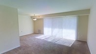 This single-story space features an open living area designed to maximize natural light and comfort. The room is anchored by a full-width sliding glass door dressed with vertical blinds, allowing ample daylight to filter in and cast soft, even light across the neutral-toned, wall-to-wall carpeting below. The walls are finished in a clean, light beige, contributing to the room's airy and understated aesthetic. A centrally positioned pendant light fixture hangs from the smooth, flat ceiling, providing a focal point and supplemental overhead lighting. A wall-mounted HVAC vent is visibly integrated beneath the large window assembly, ensuring climate control throughout the space. The overall layout feels open and versatile, offering a blank canvas suitable for a variety of furnishing arrangements. The combination of abundant natural light, neutral finishes, and straightforward design gives the space a calm, functional ambiance well-suited for everyday living.