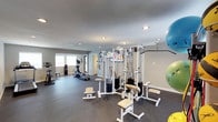 This fitness center offers a comprehensive workout environment designed to support a wide range of exercise routines. The space is finished with durable rubber flooring throughout the main workout areas, providing traction and impact absorption, while a transitional entryway features hardwood-style flooring and a seating bench beneath a large natural-light-filled window.

The cardio zone is well-appointed with a treadmill, stationary bike, and two elliptical machines, all positioned near windows that allow natural light to fill the room. The strength training area centers around a robust multi-station cable machine with multiple adjustable attachment points, accompanied by benches and a flat bench for a variety of resistance exercises.

Functional fitness accessories are also well-represented, with a dedicated rack holding an assortment of medicine balls and stability balls in multiple sizes. A large wall-mounted mirror spans one side of the strength area, enhancing the sense of space and allowing users to monitor their form. Recessed lighting throughout keeps the entire facility bright and well-illuminated. The neutral gray walls and clean white trim give the space a modern, polished aesthetic, creating a motivating and professional atmosphere for users of all fitness levels.
