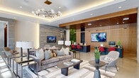 This elegantly designed single-floor space serves as a contemporary lobby and leasing center, blending modern architecture with warm, inviting interiors. The entry vestibule features polished concrete floors, a rich wood-planked ceiling, and expansive floor-to-ceiling glazing that floods the space with natural light and frames views of the surrounding streetscape and mature trees outside.

The main lounge area is anchored by a stylish seating arrangement with plush sofas, an area rug, and a distinctive branching chandelier with globe pendants set within a coffered ceiling. A textured stone-clad accent wall adds depth, while a living green wall brings a touch of nature indoors behind the staffed reception desk, which is finished in warm wood tones and outfitted with workstations and display monitors.

A sun-drenched seating alcove along the glass façade features mid-century-inspired lounge chairs bathed in natural light, creating a relaxed waiting area with panoramic outdoor views. Additional areas include a curated gallery wall displaying contemporary artwork alongside sculptural pieces, and a lounge nook centered on a double-sided fireplace beneath a wall-mounted display screen. Throughout, the design palette of concrete, natural wood, stone, and soft textiles creates a cohesive, sophisticated ambiance.