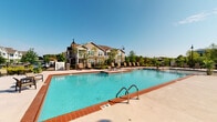 Located at 1904 Integrity Way in Louisville, Kentucky, this single-level outdoor amenity space is designed to serve as a social and recreational hub. The centerpiece is a large, sparkling swimming pool with brick-accented coping and a surrounding concrete deck lined with lounge chairs, perfect for sunbathing or unwinding beneath open skies.

An expansive covered patio extends alongside the pool area, supported by substantial columns with brick bases and a white overhead structure that provides generous shade. This sheltered space is divided into distinct functional zones. One area features a built-in outdoor kitchen with bar-height seating, offering a convenient setup for outdoor dining and grilling. Another section serves as a dedicated lounge area, furnished with a large sectional sofa with blue cushions oriented toward a wall-mounted flat-screen television, creating an inviting outdoor living room atmosphere. Brick accent walls lend a warm, grounded aesthetic throughout.

The layout flows openly between zones, with wrought iron fencing defining the pool perimeter while maintaining clear sightlines across the surrounding community. Mature landscaping and neighboring residential buildings are visible in the background, contributing to a well-integrated community feel. The space is accessible via gated entry points and is well-suited for both quiet relaxation and group gatherings.
