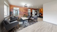 This well-appointed single-floor amenity space combines modern design with practical functionality across several distinct areas. The welcoming lounge serves as the heart of the space, featuring a striking floor-to-ceiling landscape mural in warm desert tones that anchors the room with a bold visual statement. Light wood-look tile flooring, a geometric patterned area rug, and mid-century modern seating arranged around a low-profile table create an inviting atmosphere. A sputnik-style chandelier and large windows fill the space with natural light. Adjacent to the lounge, a workstation nook with a computer provides a quiet area for focused tasks. The fitness center is thoughtfully equipped with a multi-function cable machine, a weight bench, medicine ball rack, stationary bikes, and cardio equipment, all set on durable flooring with bright overhead lighting and natural light from a window. The kitchen area is clean and contemporary, featuring white shaker-style cabinetry, a textured tile backsplash, pendant lighting, and stainless steel appliances including a dishwasher, microwave, and coffee station — ideal for communal use. Colorful artwork throughout the space adds personality and energy, tying the design together into a cohesive, modern aesthetic.