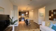 This single-floor residence showcases an open-concept layout that seamlessly connects the living and kitchen areas, creating a fluid and functional living environment. The kitchen is a standout feature, equipped with stainless steel appliances including a refrigerator, range, and microwave. Dark espresso-toned cabinetry provides strong visual contrast against light countertops, while the central island serves as both a prep space and casual dining area, accompanied by modern chrome bar stools. Elegant pendant lighting hangs above the island, adding a warm, contemporary ambiance. Rich, dark hardwood flooring runs throughout the space, grounding the design with warmth and continuity. The living area, partially visible from the kitchen, receives natural light and is accented by lush indoor greenery near the window. Neutral-toned walls keep the space feeling bright and versatile. A private entry door is visible beyond the kitchen, along with an additional interior door, suggesting additional rooms beyond the main living space. Overall, the design balances modern aesthetics with practical, everyday functionality.