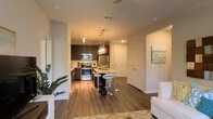 This single-floor residence showcases an open-concept layout that seamlessly connects the living and kitchen areas, creating a fluid and functional living environment. The kitchen is a standout feature, equipped with stainless steel appliances including a refrigerator, range, and microwave. Dark espresso-toned cabinetry provides strong visual contrast against light countertops, while the central island serves as both a prep space and casual dining area, accompanied by modern chrome bar stools. Elegant pendant lighting hangs above the island, adding a warm, contemporary ambiance. Rich, dark hardwood flooring runs throughout the space, grounding the design with warmth and continuity. The living area, partially visible from the kitchen, receives natural light and is accented by lush indoor greenery near the window. Neutral-toned walls keep the space feeling bright and versatile. A private entry door is visible beyond the kitchen, along with an additional interior door, suggesting additional rooms beyond the main living space. Overall, the design balances modern aesthetics with practical, everyday functionality.