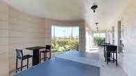 This single-level covered outdoor recreation space offers a welcoming environment designed for leisure and social activity. A prominent ping-pong table serves as the centerpiece of the space, providing a dedicated area for active entertainment. The space features a distinctive curved architectural alcove with open-air windows that frame picturesque views of the lush, landscaped grounds beyond, allowing natural light to flood the interior and creating a seamless connection between indoors and outdoors. Bar-height tables paired with sleek black stools are positioned nearby, offering casual seating ideal for socializing or spectating. Additional outdoor dining furniture is visible along the open-air corridor, extending the usable entertaining space further. The textured ceiling is fitted with elegant pendant-style light fixtures that add a refined touch to the overall aesthetic. Surrounding greenery, including neatly trimmed hedges and mature palm trees, enhances the outdoor ambiance visible through the open walls. The space strikes a balance between architectural character and functional recreation, making it a versatile amenity well-suited for both casual gatherings and active use.