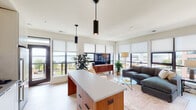 This single-floor residence showcases a thoughtfully designed open-concept layout that seamlessly connects the kitchen, dining, and living areas. Light hardwood flooring runs throughout, complementing the clean, modern aesthetic. Expansive floor-to-ceiling windows fitted with roller shades flood the interior with natural light while framing panoramic views of a nearby waterway and the surrounding urban skyline.

The kitchen is well-appointed with high-gloss white upper cabinetry, a glossy white subway tile backsplash, stainless steel appliances including a range, microwave, and refrigerator, and a generous island featuring warm wood cabinetry paired with a light gray countertop. Cylindrical pendant lights in matte black anchor the kitchen and living zones.

The open living area benefits from a wall of windows offering elevated city views, creating a bright and airy atmosphere. The primary bedroom features large windows with direct waterfront views, while an additional bedroom provides a serene, light-filled retreat. The bathroom is finished with hexagonal tile flooring, a wood-framed vanity, and a full soaking tub with subway tile surround. A private balcony accessible from the main living area completes this well-appointed residence.