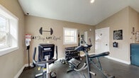 Located at 701 E Bay Dr in Largo, Florida, this dedicated fitness center offers a well-organized and fully equipped exercise space designed to support a comprehensive workout routine. The room features vaulted ceilings that lend an open, airy feel, complemented by ceiling fans and a wall-mounted mini-split unit to maintain a comfortable environment during workouts. Natural light filters in through multiple windows, brightening the warm, neutral-toned walls adorned with motivational typography decals, including the iconic "No Pain... No Gain" and "Eat. Sleep. Lift. Repeat." phrases. The dark epoxy flooring provides a durable, easy-to-maintain surface throughout. Equipment includes a treadmill, elliptical, stationary bike, and a multi-station cable machine for cardio and strength training. A full dumbbell rack with an adjustable weight bench serves free weight enthusiasts, while a medicine ball rack adds versatility. Wall-mounted flat-screen TVs on opposite ends of the room offer entertainment during sessions. A large wall mirror enhances the sense of space and allows for proper form monitoring. Additional amenities include a water dispenser, a fire extinguisher, and posted facility rules, reflecting attention to safety and user convenience. A single entry door provides straightforward access to this thoughtfully appointed fitness facility.