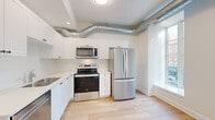 Located at 168 Wellington Street in the heart of Kingston, Ontario, this one-bedroom, one-bathroom unit offers a thoughtfully designed single-floor layout with a clean, modern aesthetic throughout. Light hardwood flooring runs continuously across the space, complementing the bright, neutral colour palette.

The open-concept kitchen and living area serves as the heart of the unit. The kitchen is outfitted with white shaker-style cabinetry, light stone countertops, and a full suite of stainless steel appliances — including a French door refrigerator, electric range, over-the-range microwave, and dishwasher. Exposed metallic ductwork traces the ceiling, lending an industrial character that contrasts tastefully with the unit's otherwise clean finishes. Large, tall windows along the exterior wall flood the living area with natural light and frame views of the vibrant Wellington Street streetscape below.

The bedroom is a well-proportioned private retreat featuring a built-in closet with a panelled double door, light flooring, and crisp white walls. The bathroom is efficiently laid out with a tiled stand-up shower, toilet, and a vanity with white cabinetry. A hallway connects the main rooms, providing a clear and functional flow throughout the unit. This space is well-suited for those seeking a modern urban residence in a central Kingston location.
