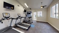 This single-story fitness center offers a comprehensive workout environment designed to support a variety of exercise routines. The main cardio area is generously sized, featuring multiple TRUE-brand treadmills, an elliptical trainer, and a stationary bike, all arranged to maximize open floor space. Durable, multi-tone carpet tile flooring covers the workout area, providing both comfort and practicality underfoot.

The space benefits from soaring vaulted ceilings that lend an open, airy feel, complemented by dual ceiling fans that ensure consistent air circulation throughout. A large bank of windows along one wall floods the interior with abundant natural light and offers pleasant views of the surrounding mature trees and landscaped grounds. An exterior door provides convenient direct access to the outdoors.

A separate adjoining room serves as an additional workout zone, equipped with a recumbent bike and stretching mats. Wall-mounted flat-screen televisions are positioned throughout for entertainment during workouts. The secondary area also includes a dumbbell rack, stability ball, and additional strength training equipment, along with a mirror wall for form monitoring. Informational signage is posted near the entrance. The overall ambiance is clean, bright, and functional, creating an inviting environment conducive to fitness.