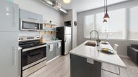 This single-floor unit showcases a thoughtfully designed open-concept layout with contemporary finishes throughout. Light wood-style flooring flows consistently across all areas, creating a cohesive and modern aesthetic.

The kitchen serves as a central highlight, featuring stainless steel appliances including a refrigerator, electric range, and microwave. Dark gray cabinetry pairs with crisp white countertops, while an island with an integrated sink and pendant lighting adds both functionality and style. Exposed ductwork above lends an industrial character to the space. Natural light enters through a large window fitted with horizontal blinds.

The living area provides a comfortable, intimate setting with track lighting overhead and additional natural light from a window. The neutral gray walls offer a versatile backdrop for a range of décor styles.

Two well-proportioned bedrooms each feature wood-style flooring and neutral tones, creating calm, restful environments. A ceiling fan is present in one room, enhancing comfort.

The bathroom is finished with a frameless glass walk-in shower with a rainfall showerhead, light-colored cabinetry, open shelving for storage, and wood-style flooring, completing the unit's clean, modern character.
