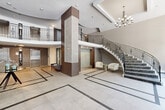 The lobby at 5200 Lakeshore Road in Burlington, Ontario, makes an immediate and lasting impression through its refined architectural design and thoughtfully curated details. At the heart of the space is a dramatic rotunda with a recessed circular ceiling that draws the eye upward, complemented by a striking sputnik-style chandelier that anchors the room with a contemporary yet timeless elegance. The expansive floor is finished in large-format neutral stone tiles accented with contrasting inlay borders, lending a sense of grandeur and order to the open space.

A sweeping curved staircase, lined with an ornate wrought iron balustrade featuring decorative scroll detailing, rises gracefully to an upper mezzanine level, which overlooks the main floor through an elegant railing. Multiple elevators, framed by richly textured mosaic tile surrounds, provide convenient vertical access throughout the building.

Floor-to-ceiling glazed windows along the entrance façade flood the lobby with abundant natural light and offer open views of the tree-lined streetscape outside. Seating areas throughout — including tufted benches, lounge chairs, and a velvet sofa — create welcoming vignettes for residents and visitors. Decorative accent walls, warm wood paneling, and carefully placed sculptural elements complete the polished, hotel-inspired ambiance of this distinguished entrance.