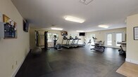 This fitness center is thoughtfully designed to accommodate a comprehensive workout experience within a single-floor layout. The main exercise area is generously sized, featuring durable dark rubber flooring that covers the majority of the workout space, transitioning to light wood-style flooring near the entry corridor and ancillary areas.

The cardio section is well-stocked with multiple treadmills, an elliptical trainer, a stationary bike, and a rowing machine, all positioned to take advantage of the natural light streaming in through several windows along the perimeter walls. Wall-mounted televisions are strategically placed throughout the space to keep users engaged during workouts.

The strength and functional training area includes a multi-station cable machine, a dumbbell rack with a full range of weights, a stability ball, and open floor space for stretching or mat work. Fluorescent ceiling fixtures provide consistent overhead lighting throughout.

The facility includes an accessible restroom, a water fountain station, a bulletin board for announcements, and built-in storage with closeted areas near the entry. French-style glass doors on multiple sides of the room connect to the exterior, enhancing ventilation and reinforcing the bright, clean ambiance of the space.