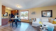 This single-floor residence is designed with a functional, open-concept layout that connects the living, dining, and kitchen areas into one cohesive space. Warm yellow-toned walls and hardwood-style flooring run throughout the main living area, creating a bright and welcoming atmosphere. A large sliding glass door and picture windows allow abundant natural light to fill the space while providing views of the surrounding outdoor greenery.

The kitchen is equipped with rich wood cabinetry, granite countertops, stainless steel appliances including a gas range, microwave, and refrigerator, and a breakfast bar with seating. The living area offers generous space for seating arrangements and is complemented by a wall-mounted air conditioning unit for climate control.

The bedroom features plush carpeting, large windows that frame outdoor views, and ample room for full-sized furniture. A wall-mounted mirror and neutral palette contribute to the room's calm, restful character.

The bathroom includes a single-sink vanity with wood cabinetry, a tub-shower combination, tile flooring, and tasteful tile surround details. Throughout the unit, the design balances practicality with a comfortable, residential aesthetic suited for everyday living.