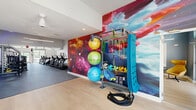 Located at 98 Northwood Blvd in Pittsboro, NC, this single-floor fitness center offers a thoughtfully designed space for a comprehensive workout experience. The main gym area commands impressive soaring ceilings with skylights, flooding the space with natural light, while bold cobalt blue accent walls with integrated LED lighting create a dynamic and energizing atmosphere. Rows of True-brand cardio machines — including treadmills, ellipticals, and stationary bikes — line the floor alongside a full complement of strength training equipment and cable machines. A vibrant, large-scale abstract mural serves as a dramatic visual focal point throughout the gym. A dedicated functional training zone features a full accessory rack stocked with stability balls in varying sizes, resistance bands, battle ropes, yoga mats, and other group fitness equipment, with direct access to the outdoors through a garage-style roll-up door. The adjoining studio room offers light hardwood flooring, floor-to-ceiling mirrors, a ballet barre, a large mounted display screen, and ceiling fans, making it ideal for yoga, barre, or group classes. A separate golf simulator room — decorated with golf-themed artwork — provides an immersive virtual golf experience. Water fountains and restroom facilities round out the amenities, ensuring a fully self-contained fitness destination.