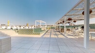 This expansive rooftop terrace offers a well-appointed outdoor retreat set against a stunning urban skyline. The space is defined by its wide-format porcelain tile flooring, which extends throughout the deck and creates a clean, modern foundation for the various amenity zones. At the heart of the terrace sits a pool with striking green and gold mosaic tile surround walls, flanked by a generous row of white lounge chairs ideal for sunbathing. A sleek white pergola cabana provides a shaded focal point beside the pool area, while an extended pergola structure runs along one side of the deck, adorned with decorative hanging lanterns and housing a series of outdoor kitchen stations equipped for entertaining. Lush ornamental grasses and potted greenery are thoughtfully integrated throughout, softening the hardscape and adding a natural element to the design. Directional signage indicates additional amenities including a dog park, games area, fire pit lounge, forum and splash pad, outdoor cabanas, and a sundeck. The rooftop commands panoramic views of the city skyline, with a vivid large-scale mural on a neighboring building adding a distinctive artistic backdrop to the outdoor experience.