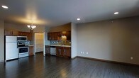 This single-floor space offers a clean, open-concept layout that seamlessly connects the kitchen and living areas, creating a sense of openness and ease of movement. The kitchen is a standout feature, showcasing rich dark-stained wood cabinetry paired with light countertops and a stylish mosaic tile backsplash that adds texture and visual interest. Stainless steel appliances, including a dishwasher, complement the modern aesthetic. Recessed lighting above the kitchen provides focused illumination, while a decorative chandelier near the entryway adds a warm, inviting touch. The open living area beyond the kitchen offers generous space, defined by neutral gray walls that provide a versatile backdrop for a variety of interior styles. Dark hardwood flooring flows consistently throughout, grounding the space with warmth and elegance. A doorway leads to an adjacent area, suggesting additional rooms or storage. The overall ambiance is modern yet welcoming, with a neutral color palette and quality finishes that make this space both functional and visually appealing.