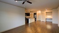 This single-floor space is designed around an open-concept layout that connects the living area and kitchen in one continuous, flowing space. The flooring throughout is a warm hardwood-style material that adds cohesion and visual warmth across the entire area.

The kitchen is a standout feature, equipped with dark espresso-toned cabinetry that contrasts sharply against the light countertops. A central island provides additional prep space and serves as a natural dividing point between the kitchen and living area. Stainless steel appliances, including a refrigerator, microwave, and range, are integrated seamlessly into the cabinetry. Pendant lighting hangs above the island, complementing the track lighting installed along the kitchen ceiling.

The living area is open and spacious, with a ceiling fan overhead providing both function and an additional design element. The neutral, cream-toned walls keep the space bright and versatile. A hallway visible in the background leads to additional rooms, with two paneled doors suggesting private spaces such as bedrooms or storage. Overall, the space presents a clean, modern, and functional design.