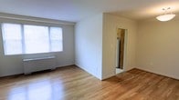This single-floor space offers a clean and functional layout highlighted by warm hardwood flooring that runs throughout the main living area. Large windows fitted with horizontal blinds allow natural light to fill the room, creating a bright and welcoming atmosphere. A baseboard heating unit beneath the windows ensures comfort year-round. The walls are finished in a neutral white tone, providing a versatile backdrop suitable for a variety of interior styles. The open living area is well-proportioned, offering ample room for furnishing and personalization. A partial wall divides the main space, leading to an adjoining area that provides access to the kitchen, where a refrigerator is visible. The kitchen appears compact and efficient, thoughtfully connected to the main living space for ease of use. The overall design is straightforward and practical, with clean lines and minimal ornamentation throughout. The combination of natural light, hardwood flooring, and a simple, open layout gives the space a bright, airy quality that makes it feel comfortable and livable.