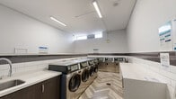 Located at 9502 Erickson Dr in Burnaby, BC, this shared laundry facility is a thoughtfully designed, single-floor space that offers a clean and functional environment for residents. The room accommodates a generous row of commercial-grade stainless steel front-loading washing machines and dryers, each equipped with digital displays for ease of use. The walls are finished with a classic white subway tile wainscoting accented by a horizontal band of dark grey tile, lending the space a clean, modern aesthetic. Above the tile, the walls are painted in a neutral light grey, keeping the environment bright and open. Flooring throughout features a striking herringbone-pattern wood-look tile that adds a stylish touch uncommon in utility spaces. A long white folding table with stainless steel legs provides ample surface area for sorting and folding laundry. Additional built-in countertop space with dark cabinetry offers further convenience. Overhead, linear LED lighting ensures the room is well-lit, while high clerestory windows along one wall allow natural light to filter in. An information board near the entrance displays relevant notices and usage instructions. The space is well-maintained, efficiently organized, and designed to serve residents comfortably.
