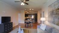 This single-floor space is designed with an open-concept layout that seamlessly connects the living room, dining area, and kitchen, creating a cohesive and functional environment. The living area is outfitted with plush seating, decorative accent pillows, and a patterned area rug that anchors the space, while a ceiling fan overhead provides both comfort and visual appeal.

Rich hardwood flooring extends throughout, adding warmth and continuity across the open floor plan. The dining area is centrally positioned, featuring a modern table with upholstered chairs, comfortably accommodating multiple guests. Track lighting mounted along the ceiling illuminates the dining and kitchen zones effectively.

The kitchen is appointed with dark cabinetry that contrasts cleanly against the lighter walls, stainless steel appliances including a refrigerator, range, and microwave, as well as ample counter and storage space. A raised breakfast bar with barstool seating provides an additional casual dining option.

Crown molding trims the ceilings throughout, contributing a refined architectural detail. The neutral wall tones keep the space feeling bright and open, making it both welcoming and versatile.