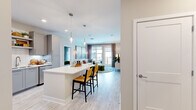 This model unit in Annapolis, Maryland offers a thoughtfully designed single-floor layout with two bedrooms, two bathrooms, and three closets. The open-concept kitchen and living area serves as the heart of the unit, featuring light wood-style flooring that flows seamlessly throughout. The kitchen is outfitted with gray shaker-style cabinetry, stainless steel appliances, a herringbone tile backsplash, open shelving, white quartz countertops, and a central island with bar seating illuminated by pendant lighting. The living area extends toward a glass door that opens to a private balcony, allowing abundant natural light to fill the space.

The primary bedroom is generously sized, featuring carpet flooring, a ceiling fan, large windows, and access to a walk-in closet with built-in shelving. The en-suite bathroom includes a glass-enclosed shower with marble-style tile surround and a single vanity. The second bedroom is similarly well-lit with large windows and carpet flooring. The second bathroom features a soaking tub with a marble-style tile surround, a gray vanity, and wood-look tile flooring. The overall aesthetic is clean, modern, and cohesive throughout.